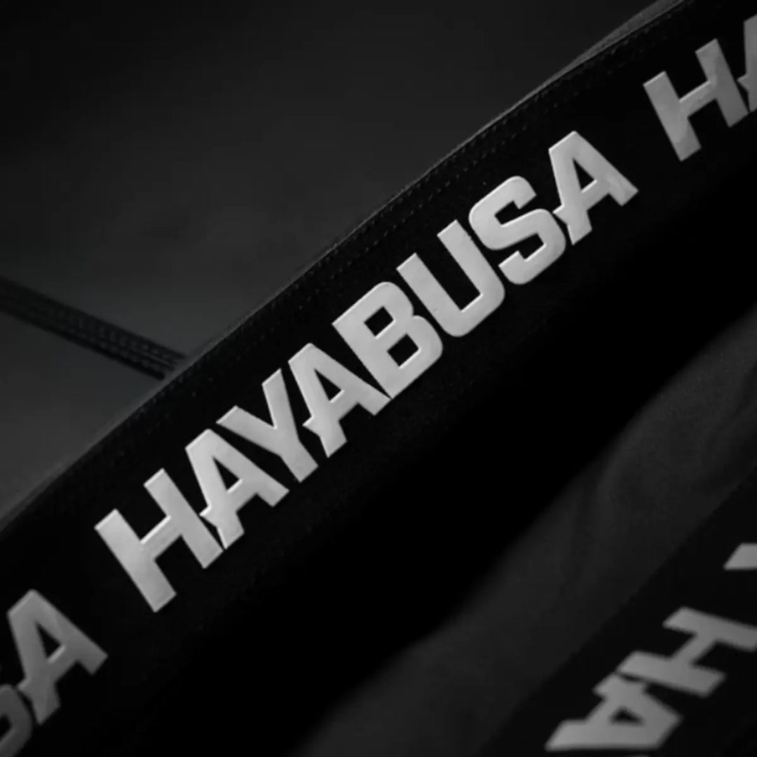 Detailfoto van de elastische tailleband met grote witte HAYABUSA-letters. De band toont stevige ondersteuning en premium afwerking. De donkere achtergrond laat het reliëf van het logo duidelijk opvallen. De afbeelding benadrukt de grip en stabiliteit van de taille.