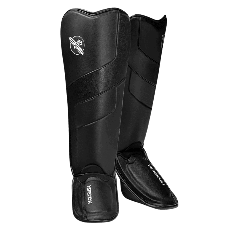 Een paar zwarte scheenbeschermers met wreefbeschermers, staand tegen een donkergroene achtergrond. De witte Hayabusa logo's zijn zichtbaar.