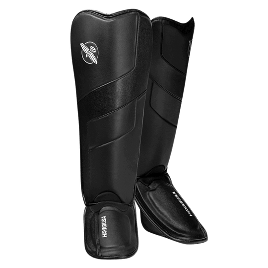 Een paar zwarte scheenbeschermers met wreefbeschermers, staand tegen een donkergroene achtergrond. De witte Hayabusa logo's zijn zichtbaar.