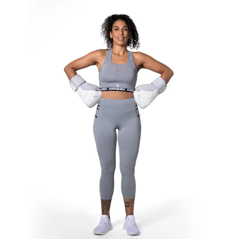 Full body view of a female athlete wearing a grey Hayabusa sports bra and matching leggings, paired with white boxing gloves and training shoes. The fitted design and neutral tone highlight a supportive, performance-focused outfit for boxing and intensive gym training.