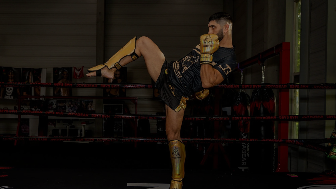 Sfeervolle actiefoto van een kickbokser die een kniestoot uitvoert in de gym, gekleed in een complete kickboks set. De sporter draagt goudkleurige bokshandschoenen en scheenbeschermers, gecombineerd met een zwart-gouden outfit. De afbeelding straalt kracht, focus en intensiteit uit en laat de bescherming en bewegingsvrijheid van de uitrusting duidelijk zien.