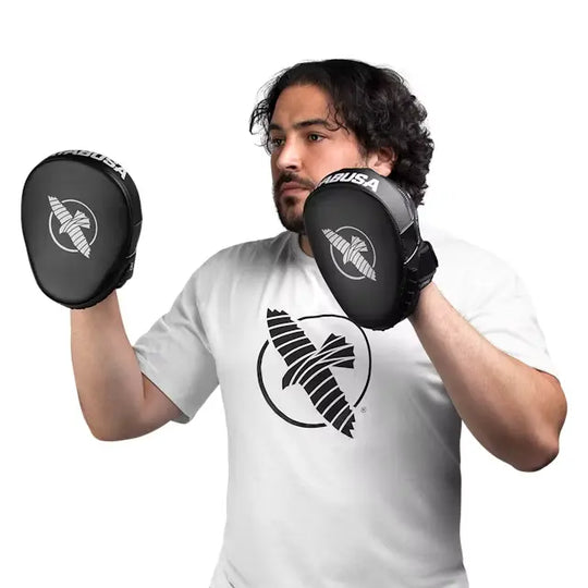 Man in een witte T-shirt met een zwart logo, die zwarte Hayabusa pads vasthoudt in een vechthouding, klaar om te trainen.