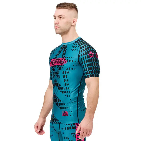 Schuin vooraanzicht van een model dat het Yokkao compression shirt Stargame in de kleur Ottanium draagt. Het turquoise shirt met zwart honingraatpatroon en roze Yokkao-logo sluit strak aan en volgt de natuurlijke lichaamsvorm. De elastische stof en korte mouwen bieden optimale ondersteuning en bewegingsvrijheid tijdens intensieve trainingen.