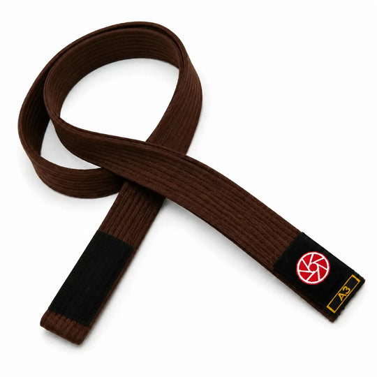 Bruine BJJ band in maat A3 met zwarte uiteinden en een rood-wit logo op het label. De stevige geweven structuur maakt de band geschikt voor BJJ-trainingen, graduaties en dagelijks gebruik op de mat.
