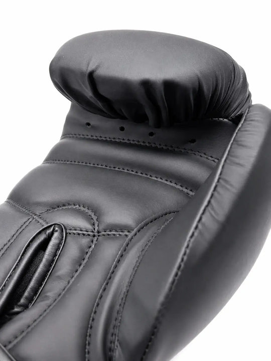 Close-up van een zwarte bokshandschoen met zichtbare stiknaden, duimbescherming en ventilatiegaatjes bij de handpalm. De stevige padding en gladde buitenlaag bieden bescherming en comfort tijdens boksen, kickboksen en intensieve stoottrainingen.