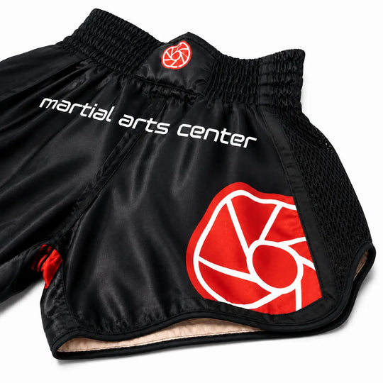 Zwart kickboksbroekje met brede elastische tailleband, meshpanelen en een opvallend rood-wit Martial Arts Center logo. De glanzende stof en ruime pasvorm maken het broekje geschikt voor kickboksen, Muay Thai en intensieve trainingen.