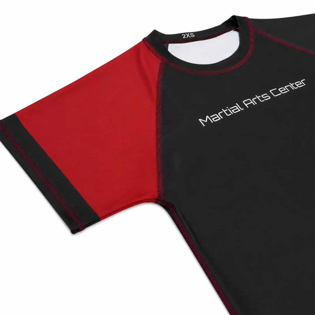 Rood-zwarte rashguard met korte mouwen, contrasterende stiksels en Martial Arts Center logo op de borst. De aansluitende pasvorm en gladde sportstof maken het shirt geschikt voor BJJ, grappling, MMA en intensieve trainingen.