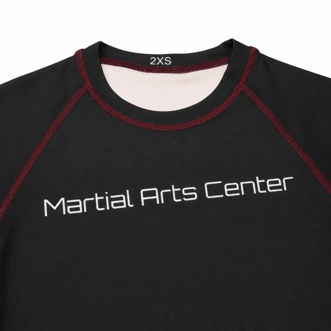 Close-up van een zwarte rashguard met Martial Arts Center logo op de borst en rode contrasterende stiksels. De aansluitende hals en gladde sportstof maken het shirt geschikt voor BJJ, grappling, MMA en intensieve trainingen.