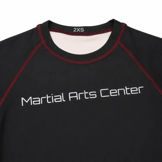 Close-up van een zwarte rashguard met Martial Arts Center logo op de borst en rode contrasterende stiksels. De aansluitende hals en gladde sportstof maken het shirt geschikt voor BJJ, grappling, MMA en intensieve trainingen.