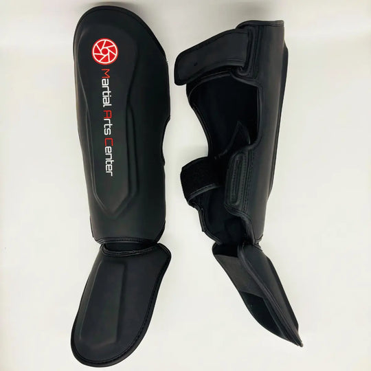 Zwarte Martial Arts Center scheenbeschermers met wreefbeschermer, wit logo en rode cirkel op de voorkant, liggend op een witte achtergrond.