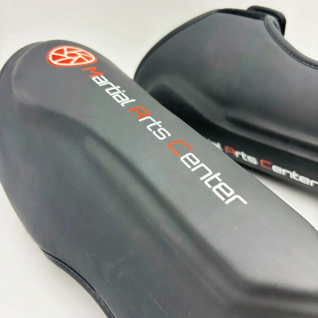 Close-up van zwarte Martial Arts Center scheenbeschermer met wit-rood logo en “Martial Arts Center” tekst op de voorkant.