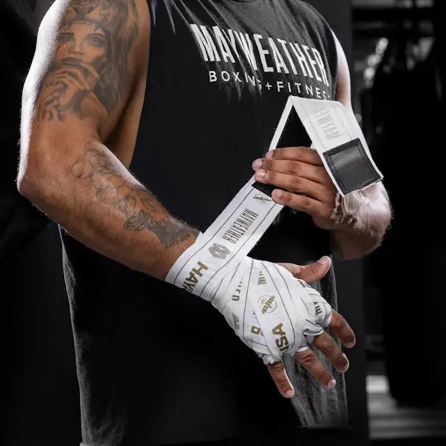 Man met tattoo wikkelt witte Mayweather Hayabusa handwraps met klittenbandsluiting om zijn handen in een boksgym, klaar voor training