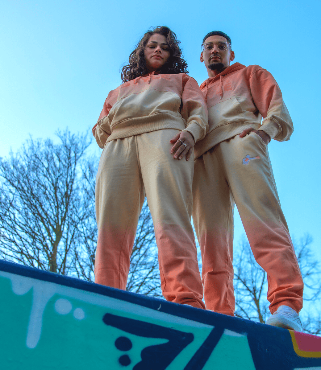 Een man en vrouw poseren samen in bijpassende oranje en crèmekleurige trainingspakken met een kleurverloop (ombré). Ze staan op een muur met graffiti, van onderaf gefotografeerd, waardoor ze dominant overkomen. Op de achtergrond is een heldere blauwe lucht met kale bomen te zien.