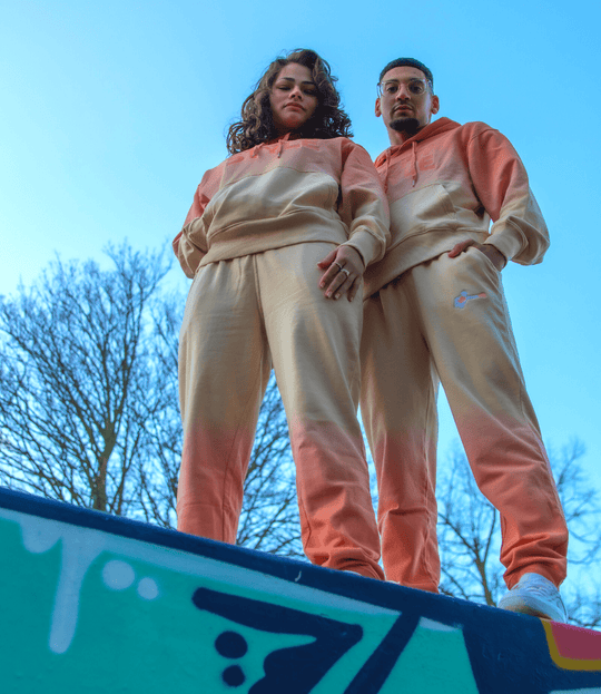 Een man en vrouw poseren samen in bijpassende oranje en crèmekleurige trainingspakken met een kleurverloop (ombré). Ze staan op een muur met graffiti, van onderaf gefotografeerd, waardoor ze dominant overkomen. Op de achtergrond is een heldere blauwe lucht met kale bomen te zien.