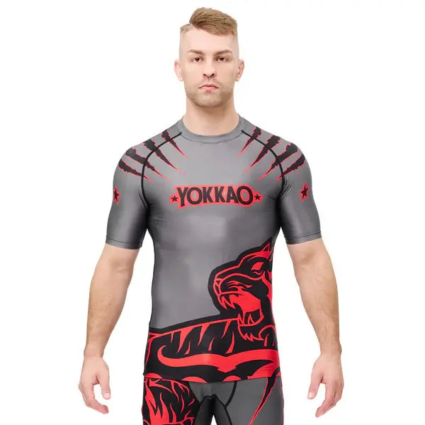 Vooraanzicht van een model dat het Yokkao compression shirt in grijs met rood en zwart design draagt. Het shirt heeft een opvallende tijgerprint aan de onderzijde en rode krasdetails op de schouders, met het Yokkao-logo op de borst. De strakke pasvorm ondersteunt het bovenlichaam en biedt maximale bewegingsvrijheid tijdens intensieve trainingen.