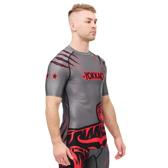 Zijaanzicht van een model dat het Yokkao compression shirt in grijs met rood en zwart design draagt. De nauwsluitende pasvorm volgt de natuurlijke lichaamsvorm en benadrukt de sportieve uitstraling. De rode details op de schouders en de tijgerprint aan de onderzijde geven het shirt een krachtige, dynamische look tijdens intensieve trainingen.