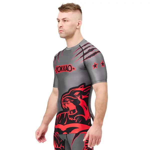 Schuin vooraanzicht van een model dat het Yokkao compression shirt in grijs met rood en zwart design draagt. De nauwsluitende pasvorm accentueert het bovenlichaam, terwijl de rode tijgerprint en krasdetails een krachtige uitstraling geven. De elastische stof en korte mouwen zorgen voor optimale ondersteuning en bewegingsvrijheid tijdens intensieve trainingen.