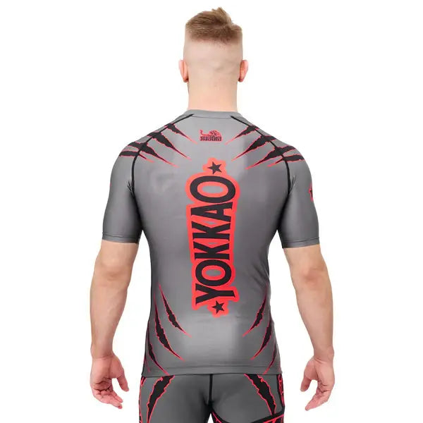 Achteraanzicht van een model dat het Yokkao compression shirt in grijs met rood en zwart design draagt. Het grote verticale Yokkao-logo in rood met zwarte omlijning domineert de rug, gecombineerd met rode krasdetails op schouders en onderrug. De strakke pasvorm ondersteunt de spieren en biedt maximale bewegingsvrijheid tijdens intensieve trainingen.