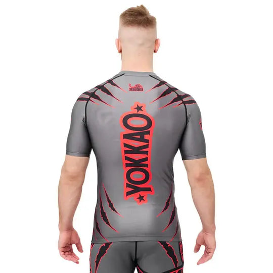 Achteraanzicht van een model dat het Yokkao compression shirt in grijs met rood en zwart design draagt. Het grote verticale Yokkao-logo in rood met zwarte omlijning domineert de rug, gecombineerd met rode krasdetails op schouders en onderrug. De strakke pasvorm ondersteunt de spieren en biedt maximale bewegingsvrijheid tijdens intensieve trainingen.