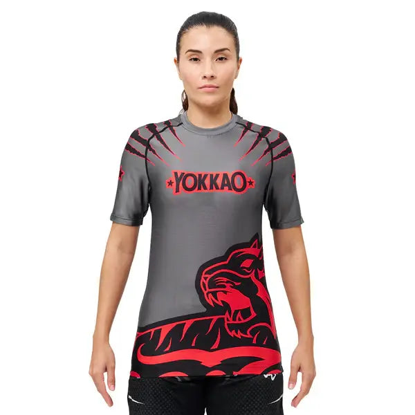 Vooraanzicht van een model dat het Yokkao compression shirt in grijs met rood en zwart design draagt. Het shirt heeft een opvallende tijgerprint aan de onderzijde en rode krasdetails op de schouders, met het Yokkao-logo centraal op de borst. De nauwsluitende pasvorm ondersteunt het bovenlichaam en biedt optimale bewegingsvrijheid tijdens intensieve trainingen.