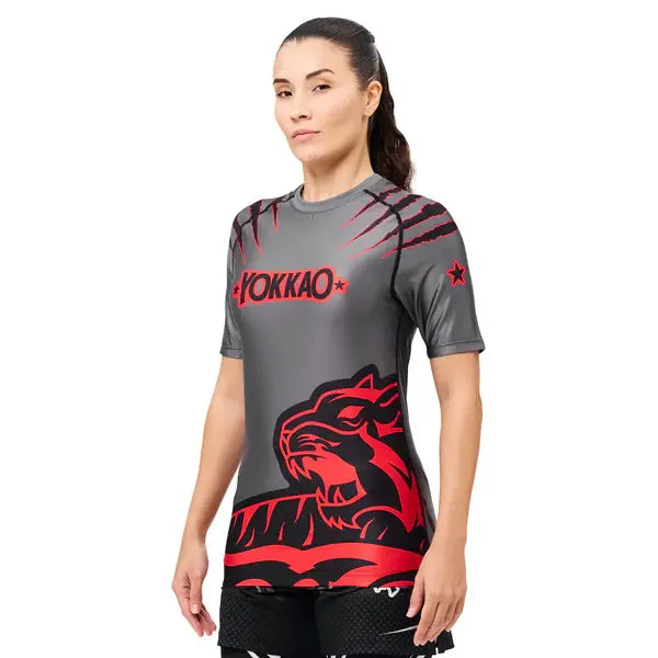 Schuin vooraanzicht van een model dat het Yokkao compression shirt in grijs met rood en zwart design draagt. De nauwsluitende pasvorm volgt de natuurlijke lichaamsvorm, terwijl de rode tijgerprint en krasdetails zorgen voor een krachtige uitstraling. De korte mouwen en elastische stof bieden optimale ondersteuning en bewegingsvrijheid tijdens intensieve trainingen.