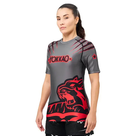 Schuin vooraanzicht van een model dat het Yokkao compression shirt in grijs met rood en zwart design draagt. De nauwsluitende pasvorm volgt de natuurlijke lichaamsvorm, terwijl de rode tijgerprint en krasdetails zorgen voor een krachtige uitstraling. De korte mouwen en elastische stof bieden optimale ondersteuning en bewegingsvrijheid tijdens intensieve trainingen.