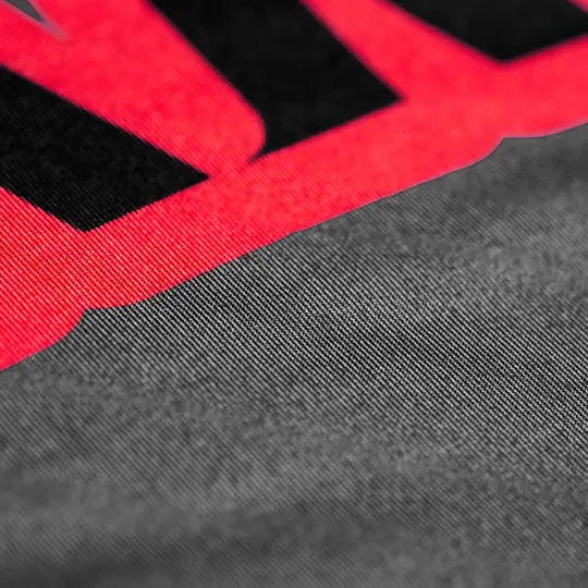 Close-up van de grijze stof en rode logo-print op het Yokkao compression shirt. De afbeelding toont de fijne structuur van het materiaal en het scherpe contrast tussen de rode en zwarte print. De gladde, elastische stof benadrukt de hoogwaardige afwerking en kwaliteit van het shirt.