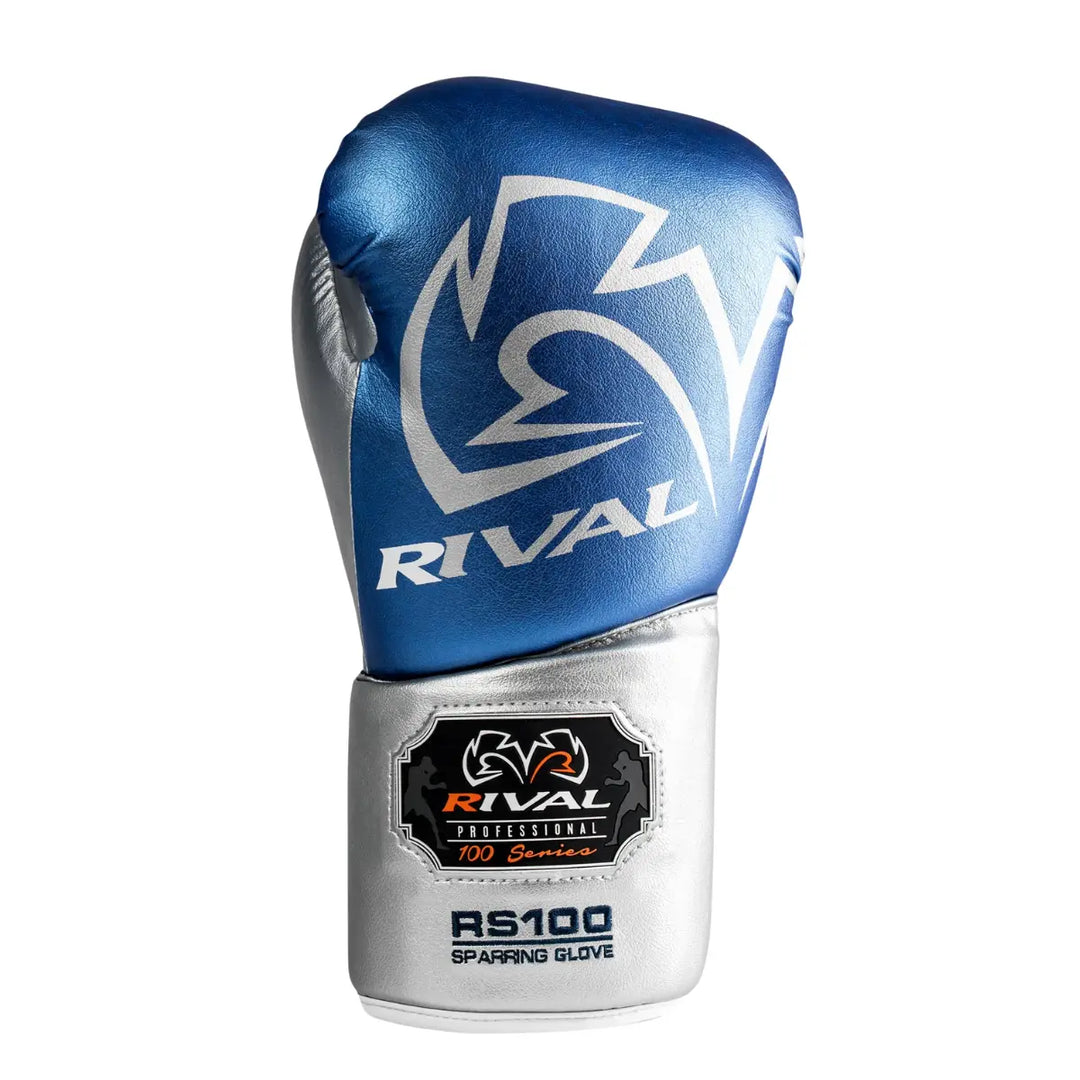 De Rival Bokshandschoen RS100 Professional Blauw Zilver is gefotografeerd op een witte achtergrond en toont een luxueus metallic design. De bovenkant heeft een glanzende blauwe afwerking met het opvallende Rival-logo in zilver, terwijl de manchet is voorzien van het RS100-embleem. De foto benadrukt de combinatie van stijl, vakmanschap en hoogwaardige bescherming die deze professionele bokshandschoen biedt.