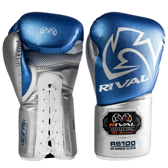 De Rival Bokshandschoenen RS100 Professional Blauw Zilver staan rechtop naast elkaar op een witte achtergrond. De ene handschoen toont de voorzijde met het opvallende Rival-logo in zilver op een blauwe basis, terwijl de andere de veterzijde en geperforeerde handpalm laat zien voor optimale ventilatie. De metallic afwerking en het RS100-embleem benadrukken het premium, professionele ontwerp van deze bokshandschoenen.