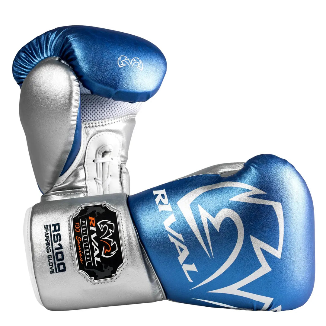 De Rival Bokshandschoenen RS100 Professional Blauw Zilver liggen gekruist op een witte achtergrond en tonen hun metallic blauwe en zilveren afwerking. Het grote Rival-logo en het RS100-embleem vallen direct op, terwijl de veterzijde en geperforeerde palm zorgen voor ventilatie en stabiliteit. Deze foto benadrukt het premium design, de ergonomische vorm en de professionele kwaliteit van deze bokshandschoenen.