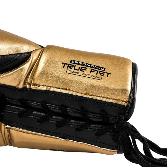 De Rival Bokshandschoen RS100 Detail Goud toont een close-up van de veterzijde van de handschoen in goud met zwarte accenten. De tekst “Ergonomic True Fist Construction” is duidelijk zichtbaar en verwijst naar het ergonomische ontwerp voor optimale pasvorm en comfort. De foto benadrukt het vakmanschap, de fijne stiksels en de hoogwaardige afwerking van deze professionele bokshandschoen.