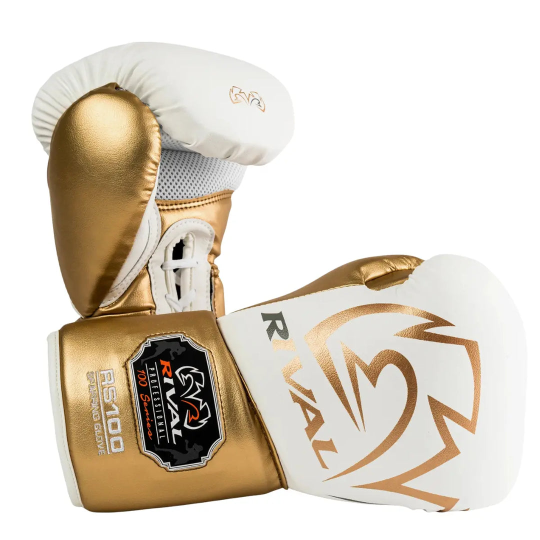 De Rival Bokshandschoenen RS100 Professional Wit Goud liggen gekruist op een witte achtergrond en tonen hun luxueuze witte en gouden afwerking. Het grote Rival-logo en het RS100 Professional-embleem vallen direct op, terwijl de veterzijde de geventileerde handpalm laat zien. De foto benadrukt het elegante design, de nauwkeurige afwerking en de professionele kwaliteit van deze high-end bokshandschoenen.