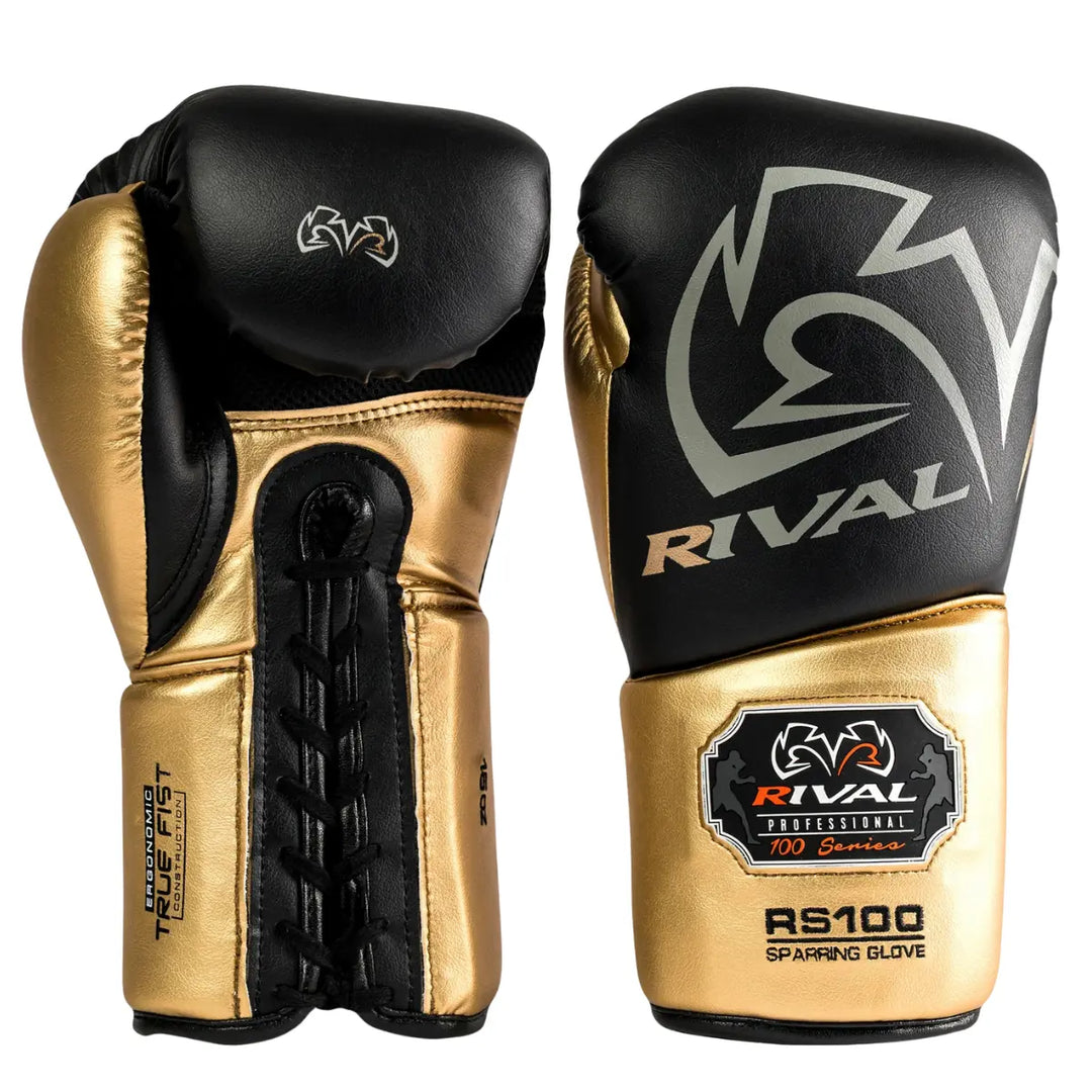 De Rival Bokshandschoenen RS100 Professional Zwart Goud staan rechtop naast elkaar op een witte achtergrond. De ene handschoen toont de voorzijde met het Rival-logo in goud, terwijl de andere de veterzijde laat zien met het opschrift “Ergonomic True Fist Construction”. De combinatie van matzwart en metallic goud straalt luxe, kracht en professionaliteit uit, perfect voor intensieve sparringssessies.