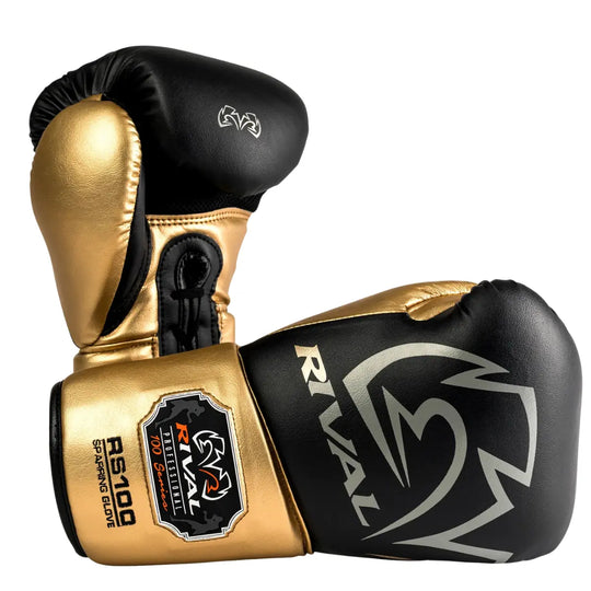 De Rival Bokshandschoenen RS100 Professional Zwart Goud liggen gekruist op een witte achtergrond en tonen een luxueus contrast tussen matzwart en glanzend goud. Het grote Rival-logo en het RS100 Professional-embleem op de manchet springen direct in het oog. Deze foto benadrukt het premium design, de professionele constructie en de exclusieve uitstraling van deze high-end bokshandschoenen.