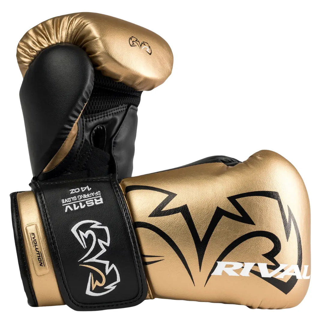 Twee Rival Bokshandschoenen RS11V Evolution Goud liggen gekruist, waarbij de ene de palmzijde toont en de andere de buitenkant met het grote zwarte Rival-logo. De combinatie van goud en zwart geeft een krachtige, luxe uitstraling. De klittenbandsluiting met “Evolution”-label en het ergonomische ontwerp benadrukken de stevigheid en het comfort van deze professionele sparringhandschoenen.