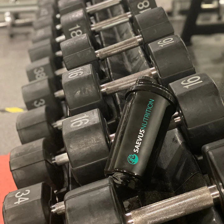Een zwarte Saevis Nutrition shakebeker geplaatst tussen een reeks dumbbells in een sportschool.