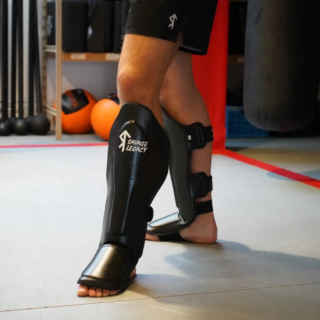 Savage Legacy scheenbeschermers in zwart gedragen door sporter in de gym, met stevige scheen- en wreefprotectie tijdens kickbokstraining.