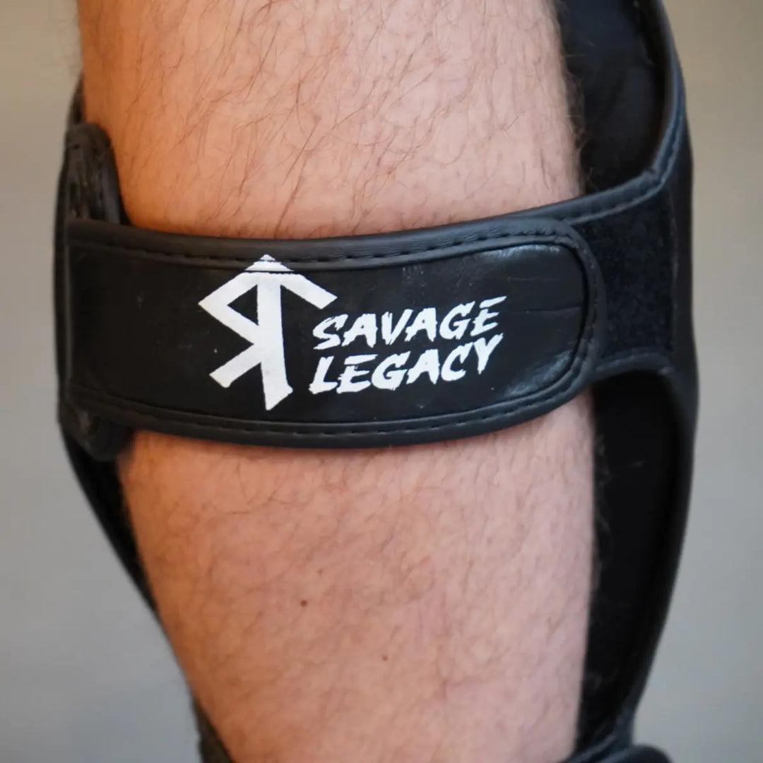 Close-up van Savage Legacy scheenbeschermer in zwart met verstelbare klittenbandsluiting en wit Savage Legacy logo rond het onderbeen.