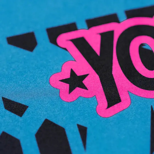 Close-up van het Yokkao-logo op het compression shirt in de kleur Ottanium. De afbeelding toont de roze en zwarte print met sterdetail op de turquoise stof. De scherpe lijnen en hoogwaardige afwerking benadrukken de kwaliteit van de print en het opvallende design.