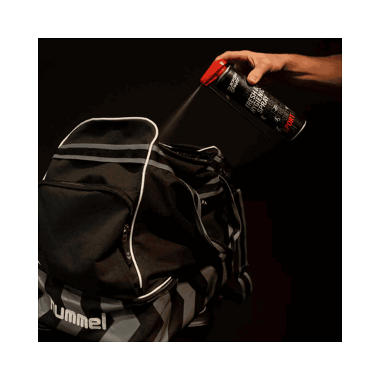 Een persoon spuit Sukkiri Spray in een sporttas om de inhoud te verfrissen en geurtjes te neutraliseren. De zwarte sporttas staat open terwijl de spray wordt aangebracht.