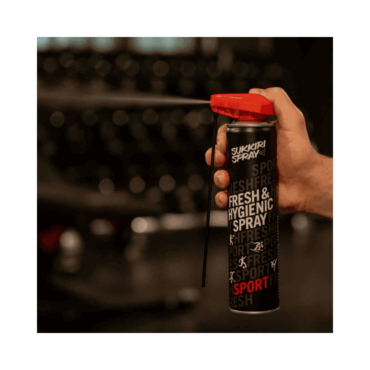 Een close-up van een hand die Sukkiri Fresh & Hygienic Spray vasthoudt en de spray activeert zonder een verlengd mondstuk. De spray is klaar voor gebruik in een sportomgeving."