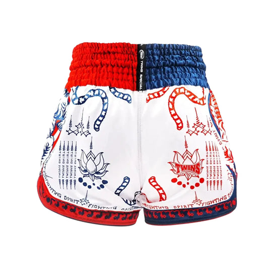 Afbeelding van de achterkant van de Twins Muay Thai short in wit met rood-blauwe details. De short heeft een tweekleurige elastische tailleband in rood en blauw met een Twins Special label in het midden. Op de achterkant staan traditionele Yant-symbolen, een lotusbloem en decoratieve patronen, met langs de onderrand de tekst “Fighting Spirit”.