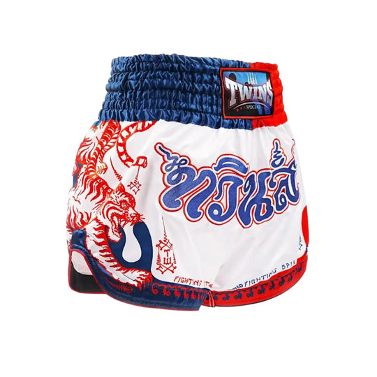 Afbeelding van een Twins Muay Thai short in wit met rood-blauwe details en traditionele Thaise Yant-print. De short heeft een brede elastische tailleband in blauw met rood accent en een Twins Special patch op de voorkant. Op de pijp staat een opvallende tijgerillustratie met Thaise tekens en decoratieve patronen langs de onderrand.