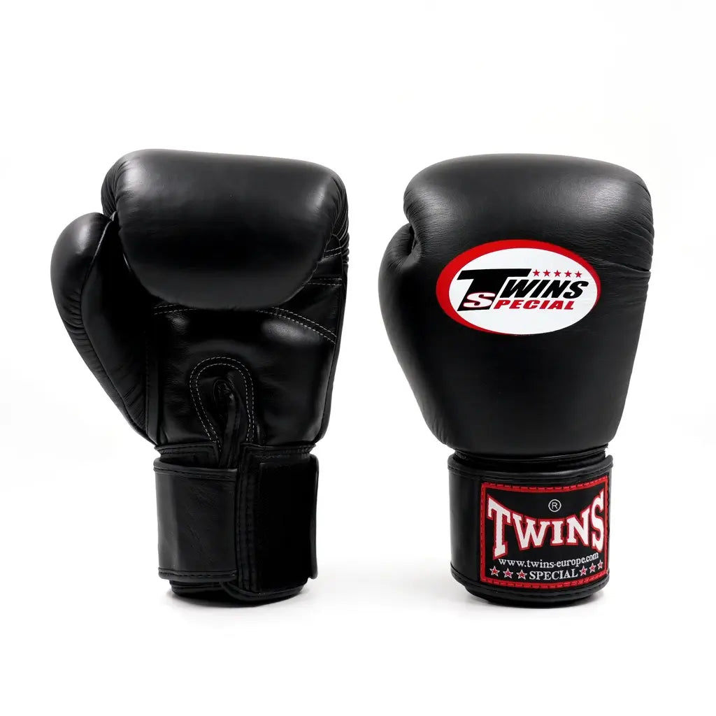 Twee zwarte Twins bokshandschoenen op witte achtergrond, met wit Twins-logo op de vuistzijde en rood label op de polsband.