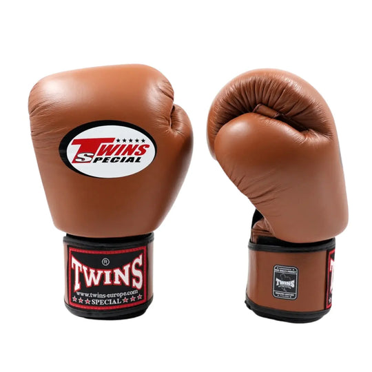 Bruine Twins Special BGVL 3 bokshandschoenen op witte achtergrond, één vooraanzicht en één zijaanzicht. Groot Twins-logo op de buitenkant en rood-zwarte manchetpatch zichtbaar. Brede klittenbandsluiting en dikke meerlagige padding voor training en sparren in boksen en kickboksen. Gemaakt van leer met een strakke, klassieke afwerking.