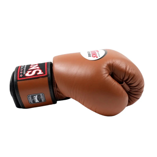 Bruine Twins Special BGVL 3 bokshandschoen in zijaanzicht, liggend met gesloten vuist. Rood-zwarte “TWINS”-manchet met brede klittenbandsluiting en ovaal Twins Special-logo zichtbaar. Dikke padding en strakke afwerking voor training en sparren in boksen en kickboksen.