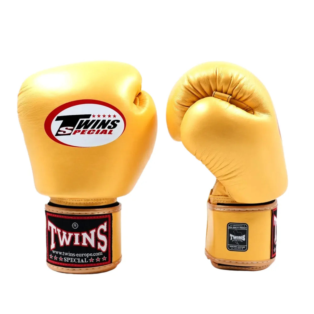 De Twins Bokshandschoenen Goud BGVL3 zijn gefotografeerd op een witte achtergrond, waarbij één handschoen rechtop staat en de andere licht gebogen is. Het Twins Special-logo is duidelijk zichtbaar op de bovenkant en op de polssluiting. De metallic gouden kleur, gladde leren structuur en stevige afwerking benadrukken de luxe uitstraling en professionele kwaliteit van deze bokshandschoenen.