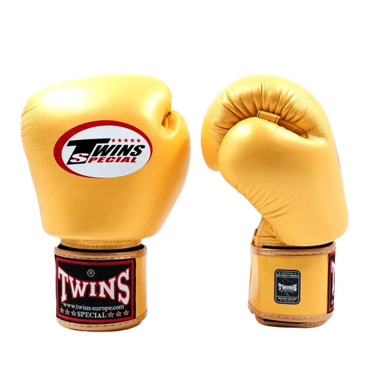 De Twins Bokshandschoenen Goud BGVL3 zijn gefotografeerd op een witte achtergrond, waarbij één handschoen rechtop staat en de andere licht gebogen is. Het Twins Special-logo is duidelijk zichtbaar op de bovenkant en op de polssluiting. De metallic gouden kleur, gladde leren structuur en stevige afwerking benadrukken de luxe uitstraling en professionele kwaliteit van deze bokshandschoenen.