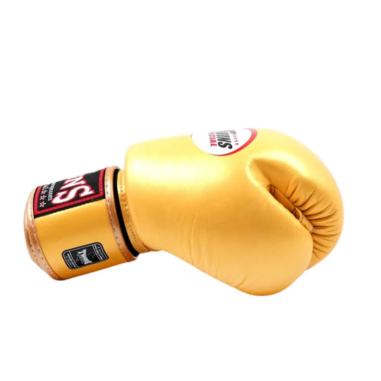 De Twins Bokshandschoen Goud BGVL3 ligt zijwaarts op een witte achtergrond met het herkenbare Twins Special-logo bovenop. De gouden kleur en glanzende leren afwerking geven de handschoen een luxueuze uitstraling. De zwarte en rode details op de polssluiting benadrukken het premium karakter en de professionele afwerking die typisch is voor Twins Special.