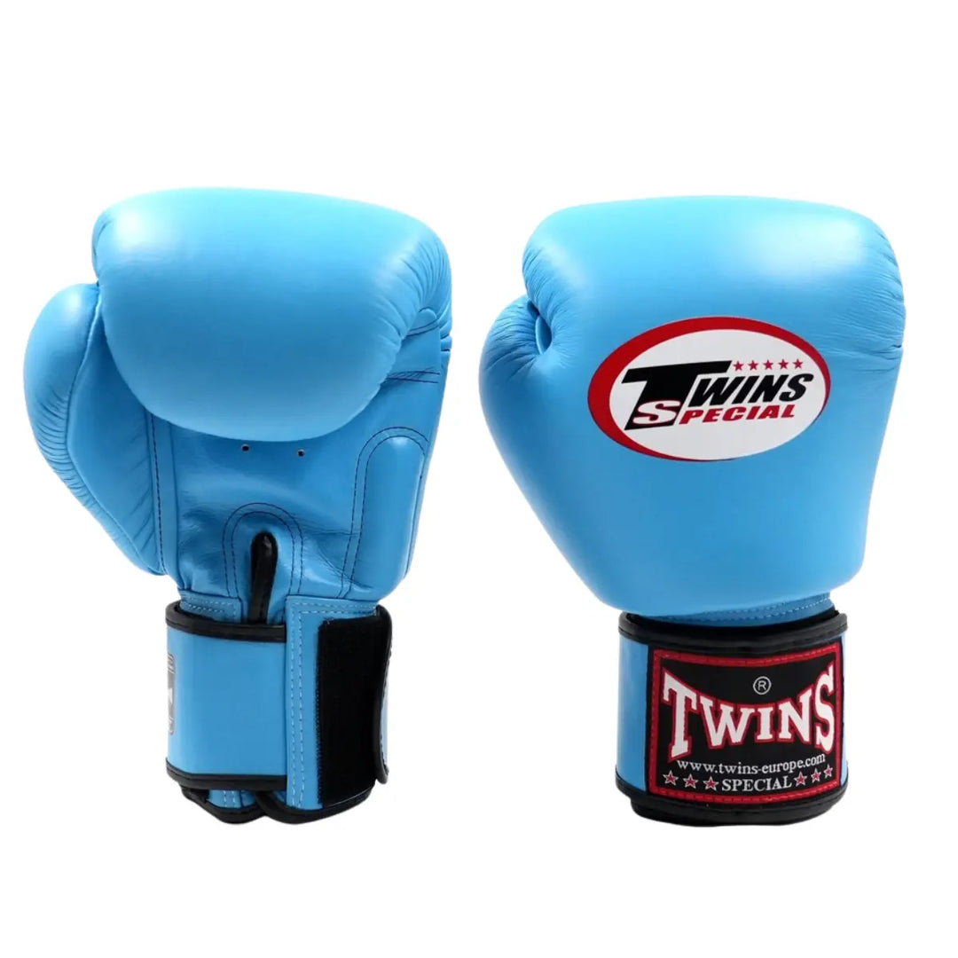 De Twins Bokshandschoenen Lichtblauw Set toont een paar lichtblauwe bokshandschoenen van Twins Special, geplaatst rechtop op een witte achtergrond. De voorkant en binnenzijde zijn zichtbaar, met het opvallende rode Twins-logo op de polssluiting en bovenop de handschoen. De gladde leren afwerking en stevige klittenbandsluiting benadrukken het professionele karakter van deze handschoenen voor intensieve bokstrainingen.
